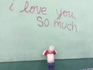 A little boy lifting up his shirt and looking at his bellybutton underneath the "I love you so much" mural in Austin, Texas